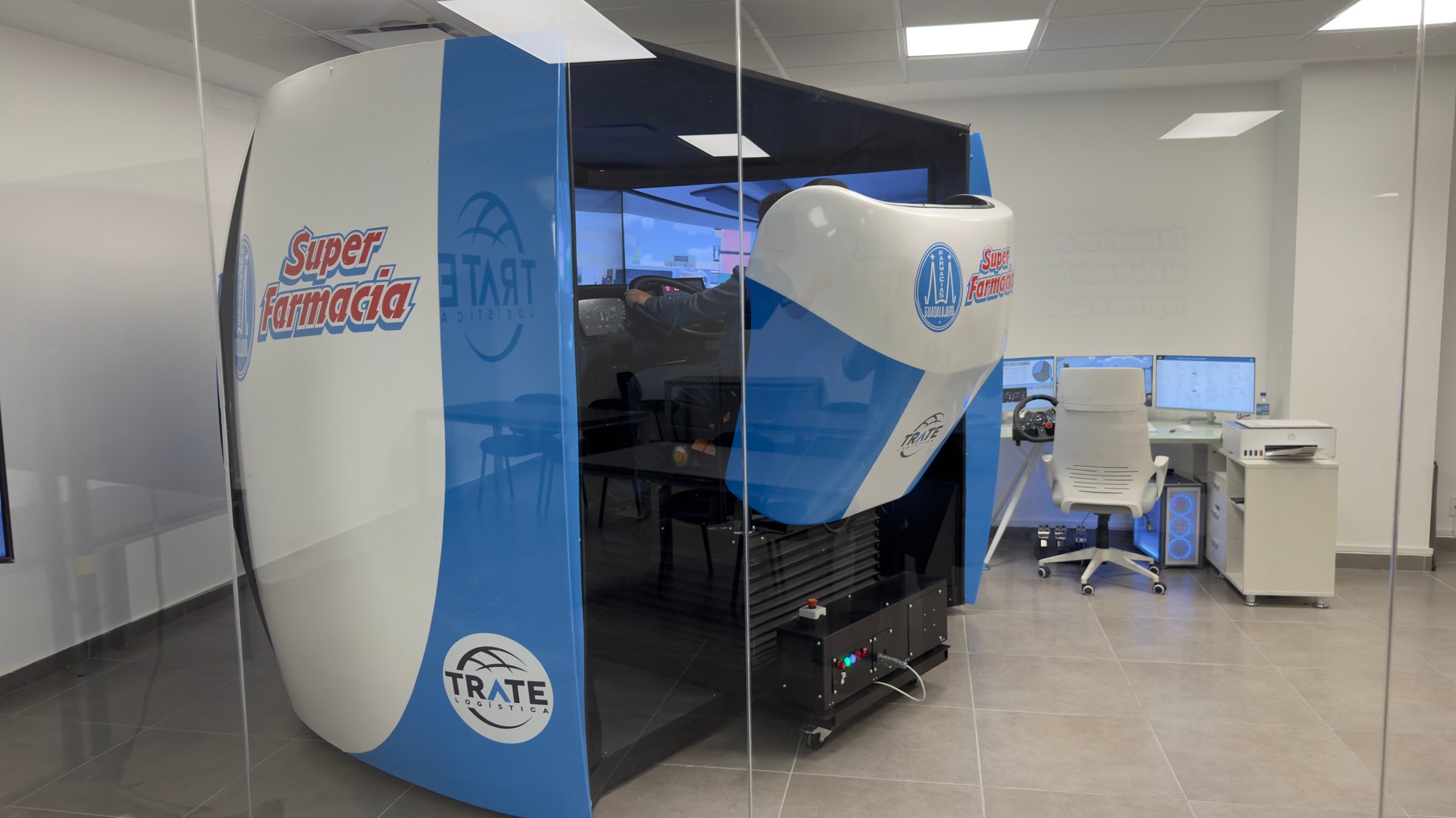 Cabina completa del simulador APEX rotulada con la identidad de Farmacias Guadalajara, junto al puesto del instructor